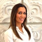 Smiling woman with long brown hair in a white blouse, standing in front of a white decorative wall relief.