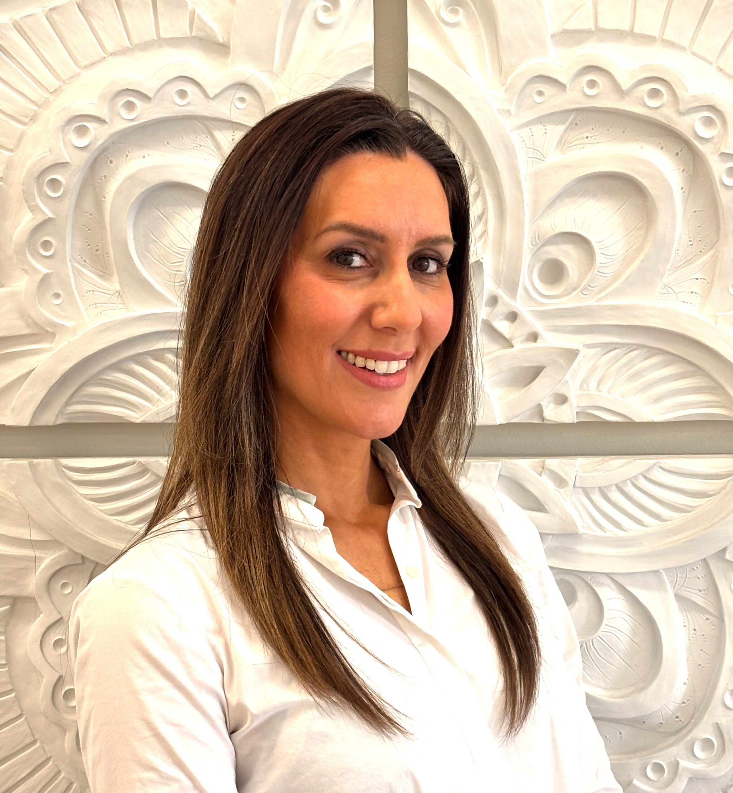 Smiling woman with long brown hair in a white blouse, standing in front of a white decorative wall relief.