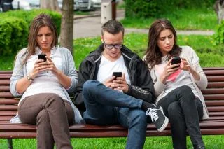 Three friends sitting on a park bench, each looking at a smartphone in their hands.